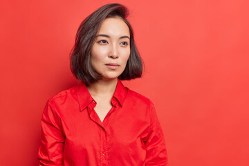 Beautiful brunette Asian woman with healthy skin has concentrated gaze into distance thinks over decision being deep in thoughts wears shirt poses against bright red background empty space aside