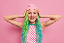 Positive European Teenage Girl Keeps Hands To Head Happy To Buy New Outfit Has Dyes Colorful Hair Has Piercing In Nose Isolated Over Pink Background. Glad Stylish Woman Shows Her Trendy Hairstyle