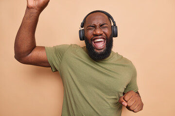 Wall Mural - Overjoyed bearded black man dances carefree keeps arms raised up moves with rhythm of music listens music via headphones dressed in casual t shirt isolated over brown background. Entertainment