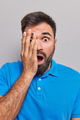 Wall Mural - Shocked frightened bearded adult man keeps palm on face drops jaw sees somethig horrible dressed in casual blue t shirt isolated over grey background. Concerned emotive guy stares in disbelief