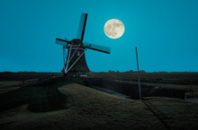 A Historic Old Grain Windmill On The North Sea Near Eemshaven. It Is Night And The Full Moon Is In The Sky. The Blue Light Illuminates The Landscape.