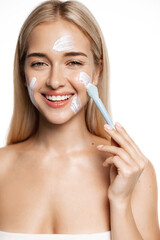 Wall Mural - Vertical shot of beautiful blond girl applying facial mask, standing in towel after shower with clean hydrated skin, using brush for lotion, face scrub, white background