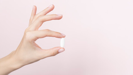 Hand holding a capsule or pill on a pink background, close up view.