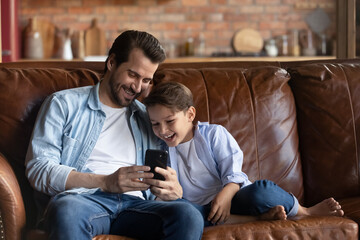 Wall Mural - Smiling loving young Caucasian father and small teen son relax on couch at home talk speak on video call on smartphone. Happy little boy child with dad rest have fun laugh use cellphone together.