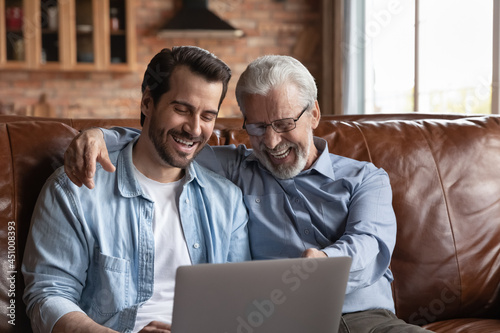 Overjoyed millennial man child relax on sofa with smiling senior father use modern laptop gadget together. Happy old dad and adult grownup son rest at home talk speak on webcam call on computer.