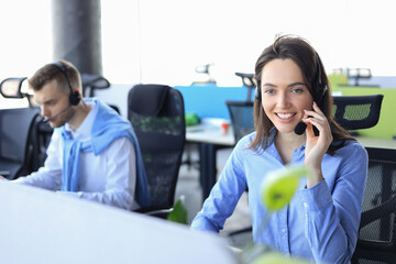 Wall Mural - Smiling male call-center operator with headphones sitting at modern office with collegues on the backgroung, consulting online.