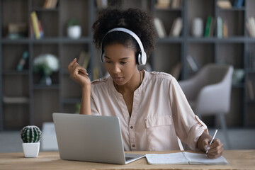 Wall Mural - Pensive millennial African American female student in earphones study online on laptop handwrite take notes. Focused young ethnic woman in headphones work distant on computer write summarize in pad.