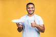Indian african american chef cook or baker man in striped apron isolated over yellow background. Cooking food concept. Mock up copy space. Hold empty blank plate with place for food.