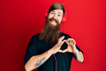 Canvas Print - Redhead man with long beard listening to music using headphones smiling in love doing heart symbol shape with hands. romantic concept.