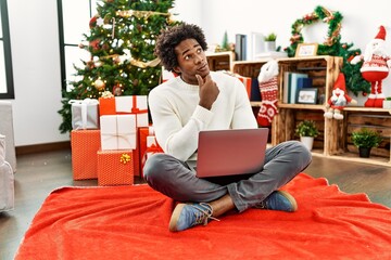 Sticker - Young african american man using laptop sitting by christmas tree serious face thinking about question with hand on chin, thoughtful about confusing idea