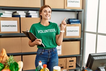 Sticker - Young brunette woman wearing volunteer t shirt at donations stand with a big smile on face, pointing with hand finger to the side looking at the camera.