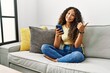 © Krakenimages.com - Beautiful hispanic woman sitting on the sofa at home using smartphone pointing to the back behind with hand and thumbs up, smiling confident