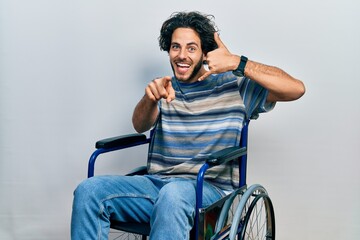 Sticker - Handsome hispanic man sitting on wheelchair smiling doing talking on the telephone gesture and pointing to you. call me.