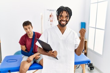 Wall Mural - Young hispanic man working at pain recovery clinic with a man with broken arm very happy and excited doing winner gesture with arms raised, smiling and screaming for success. celebration concept.