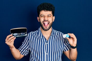 Sticker - Young arab man with beard holding glasses and contact lenses sticking tongue out happy with funny expression.