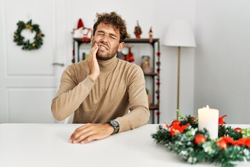 Wall Mural - Young handsome man with beard sitting on the table by christmas decoration touching mouth with hand with painful expression because of toothache or dental illness on teeth. dentist