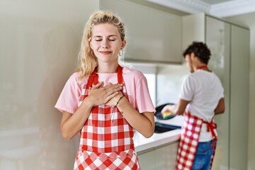 Canvas Print - Young caucasian woman wearing apron and husband doing housework washing dishes smiling with hands on chest with closed eyes and grateful gesture on face. health concept.