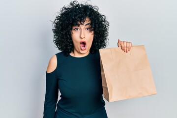 Poster - Young middle east woman holding take away paper bag scared and amazed with open mouth for surprise, disbelief face