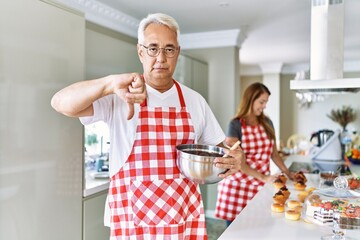 Canvas Print - Middle age hispanic couple wearing apron cooking homemade pastry with angry face, negative sign showing dislike with thumbs down, rejection concept