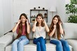 Group of three hispanic girls sitting on the sofa at home covering ears with fingers with annoyed expression for the noise of loud music. deaf concept.