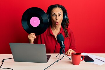 Poster - Beautiful middle age woman working at radio studio holding vinyl disc scared and amazed with open mouth for surprise, disbelief face