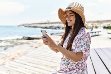 Poster - Young latin girl wearing summer using smartphone sitting on the bench hat at the beach.