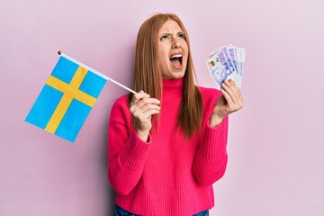 Wall Mural - Young irish woman holding sweden flag and krone banknotes angry and mad screaming frustrated and furious, shouting with anger looking up.