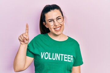 Canvas Print - Young hispanic woman wearing volunteer t shirt pointing finger up with successful idea. exited and happy. number one.