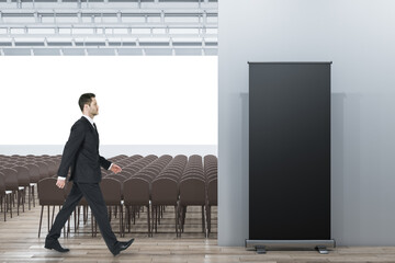 Poster - Businessman walking in contemporary auditorium interior with seating, wooden flooring and empty mock up poster on wall.