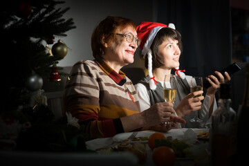 Smiling women watching Christmas tv program sitting at table with wine glasses in dark room