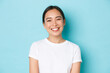 Close-up of cheerful carefree asian girl in white t-shirt smiling broadly. Pleasant korean female looking friendly at camera, enjoying nice conversation, talking with someone, light blue background