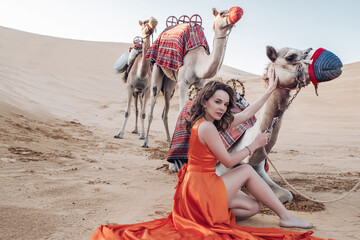 Gorgeous Caucasian woman wearing an orange long dress sitting on the sand with camels in a desert