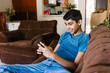 Mexican teen boy having fun with smartphone sitting on sofa at home in Latin America
