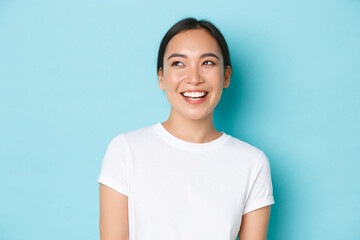 Close-up of dreamy asian girl in white t-shirt, looking hopeful and happy upper left corner, laughing and smiling broadly, standing joyful over light blue background