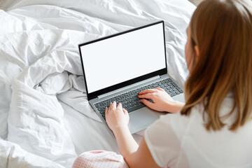 Young woman in pajamas sitting on bed using laptop With blank mockup template on screen display. Remote work from home workspace in bed using laptop with screen mock up for design. 