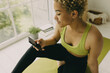 © shurkin_son - Close-up picture of delighted curly blonde short hair girl with perfect dark skin sitting in white room in fitness clothes wearing piercing typing on her phone with screen space for your advertisement