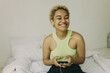 © shurkin_son - Happy, smiling African American young blonde with earphones holding phone sitting on white bed dressed in sportswear laughing while listening to audiobook at home. People and technology concept