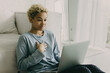 © shurkin_son - Smiling young African American woman with wild dyed blonde curly afro hairstyle at home sitting in casual clothes on floor in living room, having video chat on laptop with surprised face expression