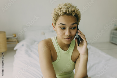 Close-up of adorable young blonde lady with perfect dark skin talking on phone with surprised face expression sitting on bed with white linen, wearing body jewelry, minimalistic interior on background