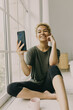 © shurkin_son - Picture of mixed race blonde girl, phone and pink coffee cup on floor near panoramic window. Curly-headed woman holding smartphone and posing for selfie, having toothy smile, touching cheek with hand