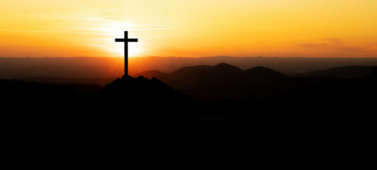 Religious grief landscape background banner panorama - View with black silhouette of mountains, hills, forest and cross / summit cross, in the evening during the sunset, with orange colored sky..