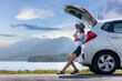 © tuaindeed - Young Asian woman traveler sitting on hatchback car trunk with mountain landscape background. Traveler car concept