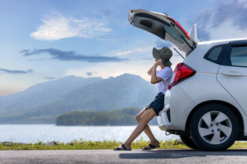 Young Asian woman traveler sitting on hatchback car trunk with mountain landscape background. Traveler car concept