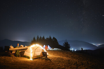 Wall Mural - Young man resting near lighted tent outdoors at night. Group of tourists relaxing on meadow under evening starry sky. Silhouettes of mountains and city lights on the background. Concept of camping.
