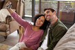 © Wavebreak Media - Happy diverse couple sitting on sofa in living room taking selfie and smiling