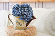 Home decor. A sprig of blue hydrangea in a decorative watering can for flowers on a round wicker straw stand.