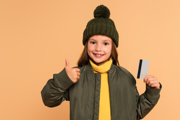 Wall Mural - happy and stylish kid showing thumb up while holding credit card isolated on beige