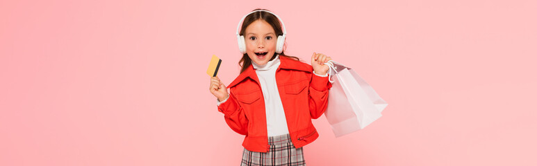 Wall Mural - excited girl in headphones and stylish outfit posing with credit card and shopping bags isolated on pink, banner