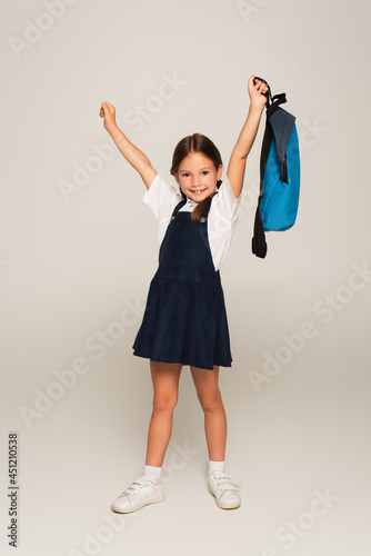 excited schoolkid with blue backpack showing win gesture on grey