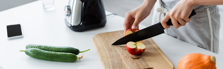 Wall Mural - cropped view of woman cutting apple near fresh cucumbers and electric blender, banner.